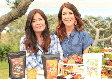 Two women seated outdoors with baking mix bags and a platter of desserts on a table.