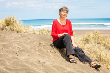 Elderly woman in red top sitting on a sandy dune by the sea with blue sky background.