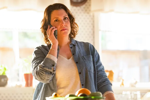 A woman in a denim shirt on the phone by a kitchen table with a fruit bowl, looking concerned.