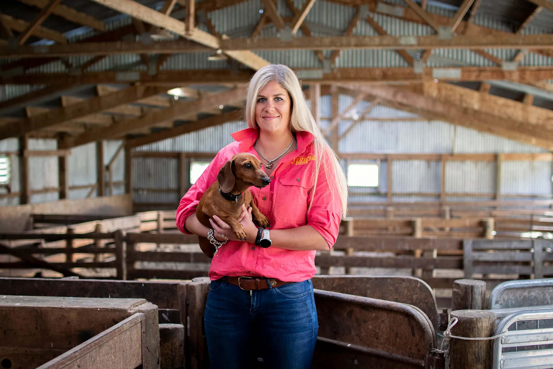 Woman in pink shirt holding a brown dachshund in a wooden barn setting.