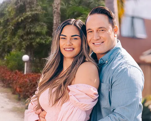 A couple smiling and embracing outdoors, with greenery in the background.