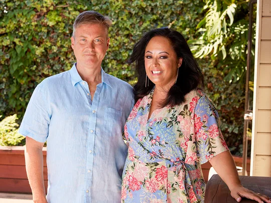 Two individuals smiling, standing outdoors in front of lush greenery, with the woman wearing a floral dress.