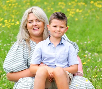 A woman and a young boy sit together on grass with yellow flowers, smiling at the camera.