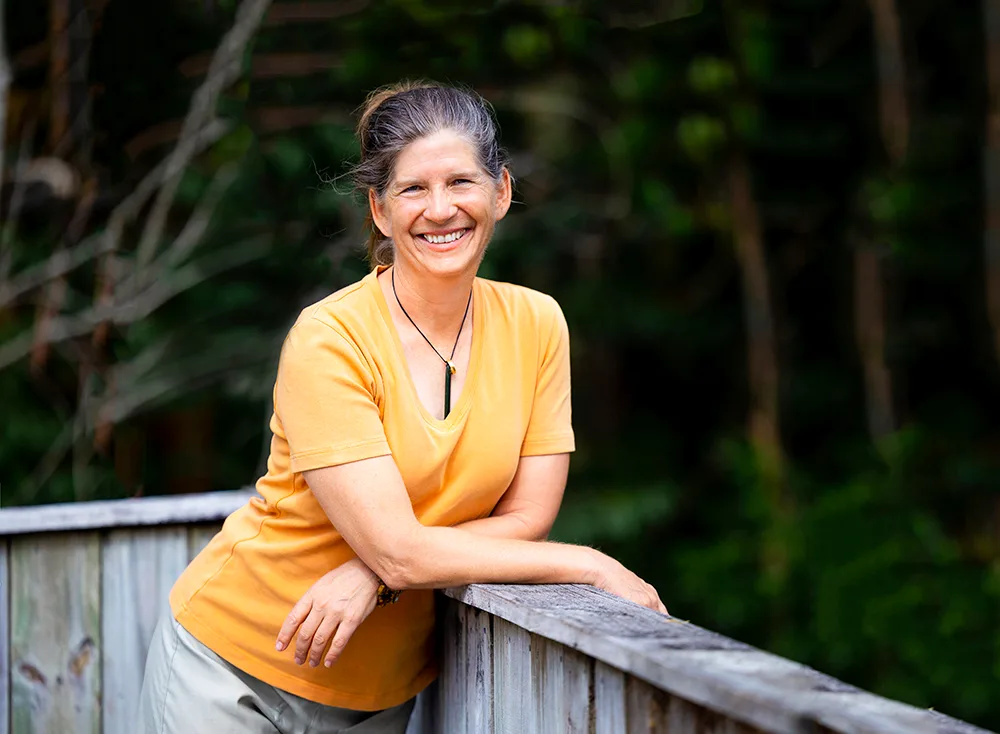 Person in an orange shirt leaning on a wooden railing, smiling in a lush green outdoor setting.