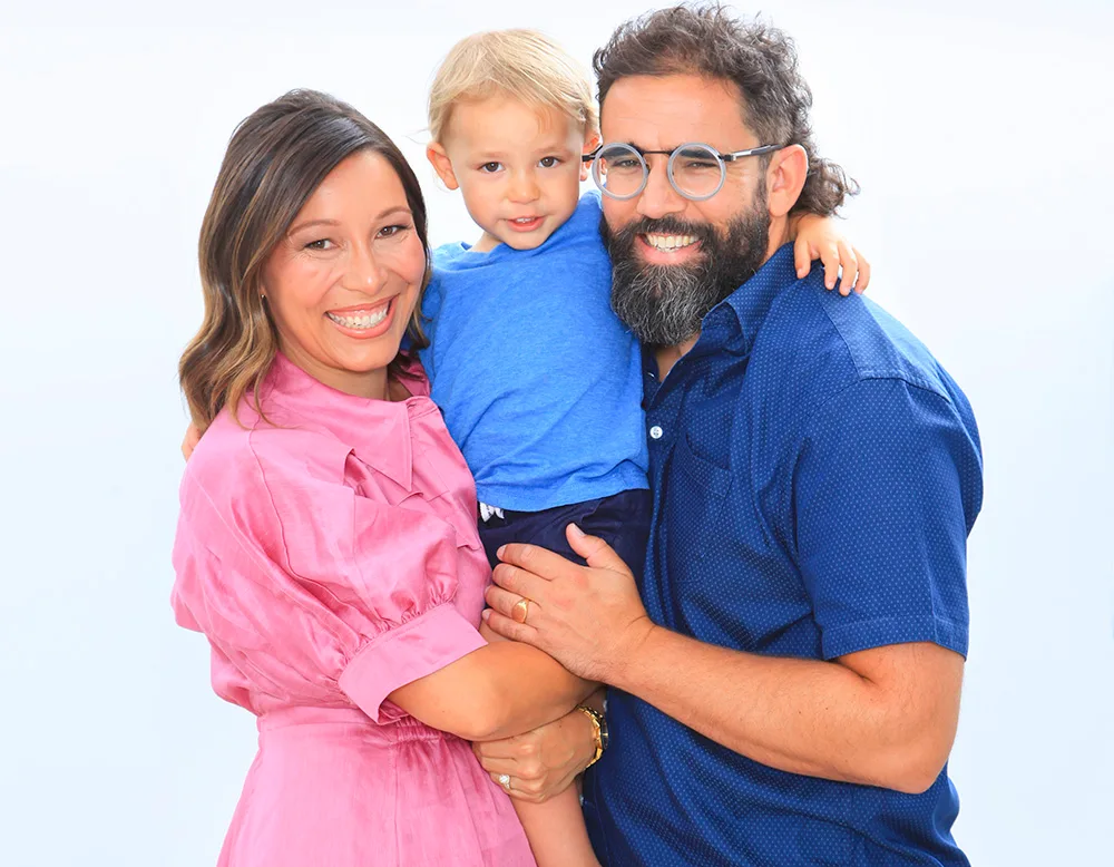 A couple smiling while holding a toddler, all dressed in vibrant colors, against a plain background.