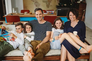 Family of five sitting on a couch, smiling. Son holds a Christmas book. World map on wall behind.