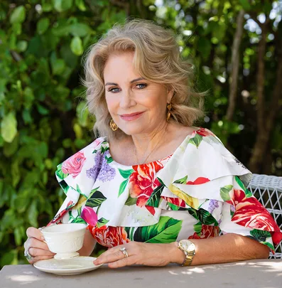 A woman sits outdoors holding a teacup, wearing a floral dress and smiling, with greenery in the background.