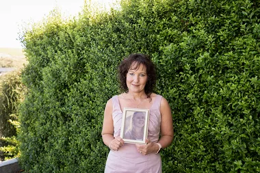 A woman in a pink dress holds a framed photo of another woman, standing in front of a green hedge.
