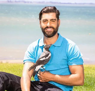 A man in a blue shirt sits by the water, smiling with a magpie perched on his arm.