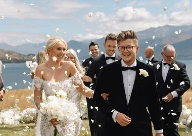 Bride and groom smiling while walking down an outdoor aisle with petals falling around them, surrounded by groomsmen.