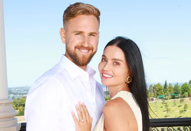 A smiling couple outdoors, embracing, with the woman showing an engagement ring.