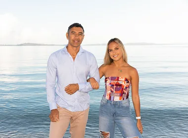 Man and woman smiling by the beach, with clear sky and calm water in the background.