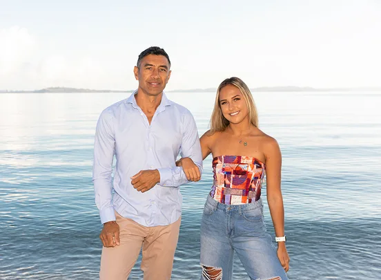 Man and woman smiling by the beach, with clear sky and calm water in the background.