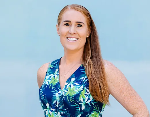 A woman with long red hair smiles, wearing a floral blue dress, and stands against a light blue background.