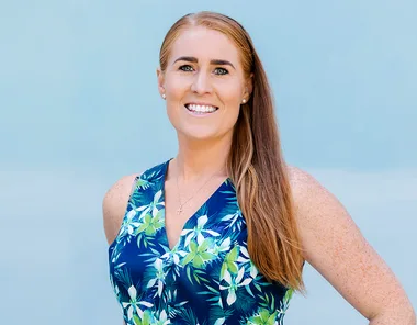 A woman with long red hair smiles, wearing a floral blue dress, and stands against a light blue background.