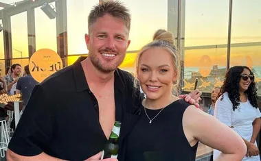 A smiling man and woman pose together at a rooftop event during sunset, with others socializing in the background.