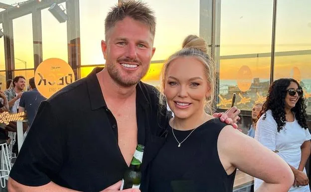 A smiling man and woman pose together at a rooftop event during sunset, with others socializing in the background.