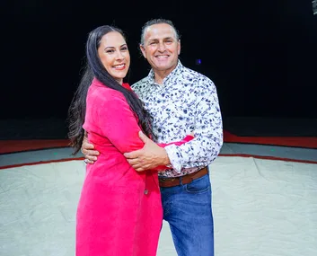 Couple smiling and embracing in the center of an indoor area, with the woman in a pink coat and the man in a patterned shirt.