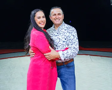 Couple smiling and embracing in the center of an indoor area, with the woman in a pink coat and the man in a patterned shirt.