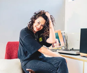 Young woman with curly hair and black shirt, sitting casually by a turntable and records, smiling warmly.