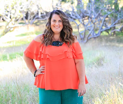 Smiling woman in an orange blouse stands in a grassy area with trees in the background.