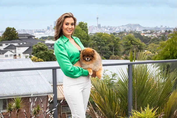 Woman in a green shirt holds a small fluffy dog on a balcony with a cityscape in the background.