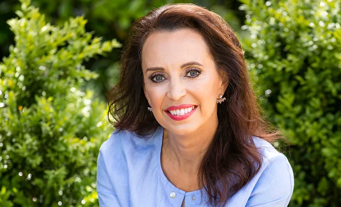 Woman with long dark hair smiling in a garden, wearing a light blue blouse.