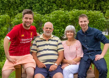 A family of four smiling, sitting on a bench in a garden, with greenery in the background.