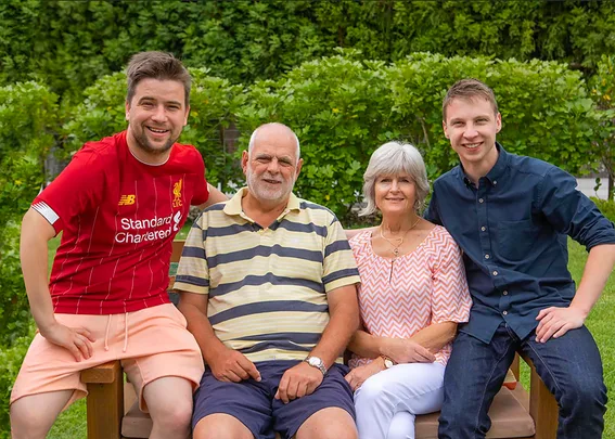 A family of four smiling, sitting on a bench in a garden, with greenery in the background.