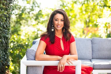 Woman in a red dress sitting on a white outdoor couch surrounded by greenery, smiling at the camera.