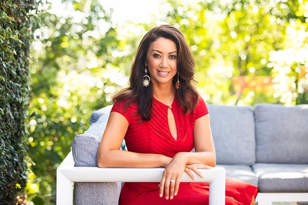 Woman in a red dress sitting on a white outdoor couch surrounded by greenery, smiling at the camera.
