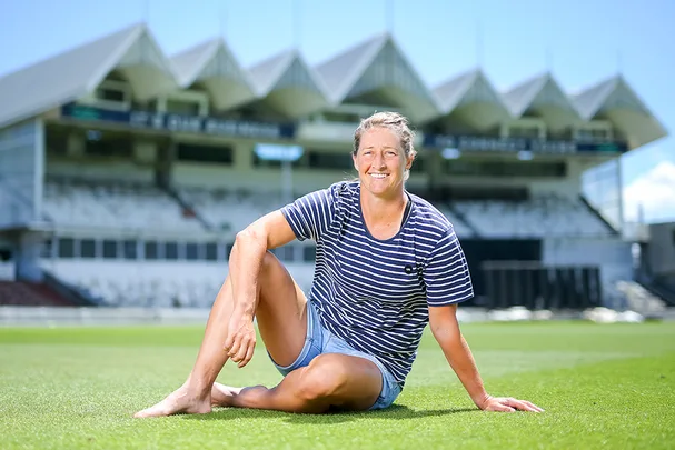 Woman in striped shirt sitting on grass with a stadium backdrop, smiling in bright daylight.