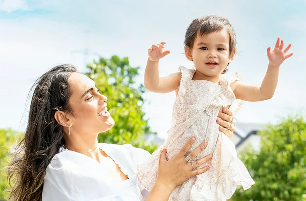 A woman lifts a smiling toddler in the air, both in white outfits, against a bright outdoor background.