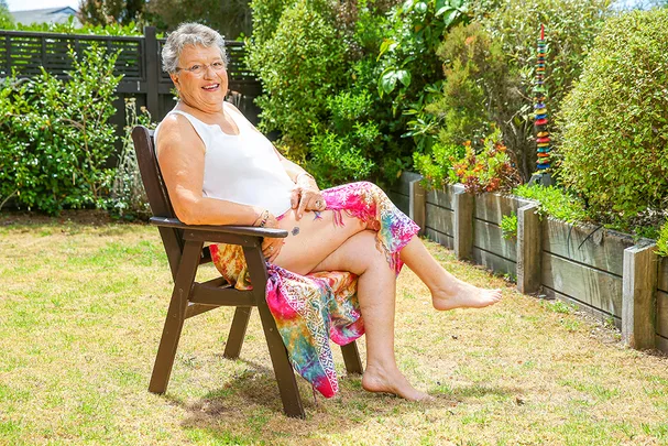 Senior woman with glasses sitting on a chair in a garden, wearing a white tank top and colorful skirt, smiling.