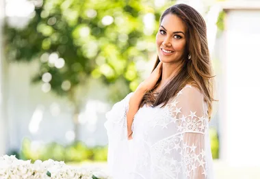 A smiling woman in a white lace outfit stands outdoors with a blurred green background.