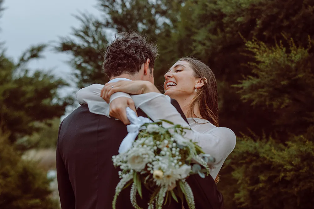 Bride embracing groom outdoors, smiling and holding a bouquet with greenery and white flowers.