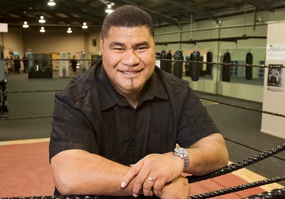 A person smiling in a boxing gym, leaning on the ring ropes, wearing a black shirt.