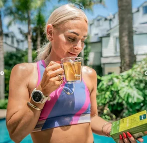 A woman in athletic attire drinks tea by a pool, holding a tea box, wearing a smartwatch, with palm trees in the background.