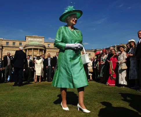 A woman in a green dress and hat, standing outside a large building, with a crowd gathered around.