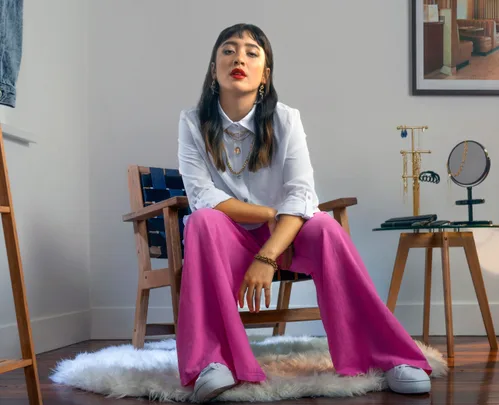 Woman in a white shirt and pink pants sits in a chair, surrounded by jewelry on a side table in a modern room.