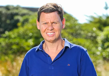 Man in a blue collared shirt standing outdoors with greenery in the background.