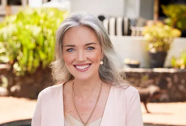 Smiling woman with long gray hair, wearing a pink blazer and gold necklace, outdoors with blurred greenery in background.