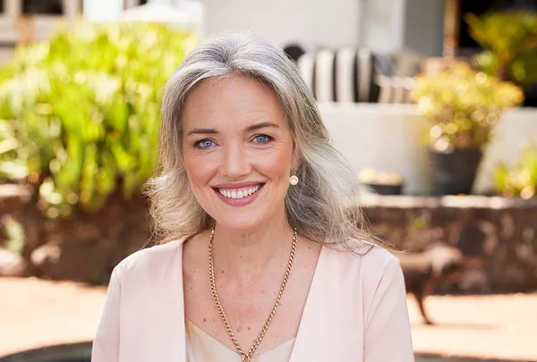 Smiling woman with long gray hair, wearing a pink blazer and gold necklace, outdoors with blurred greenery in background.