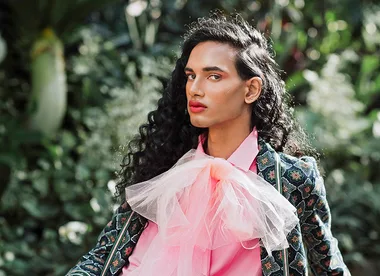 A person with long curly hair wears a floral suit and pink bow, posed against a lush greenery background.