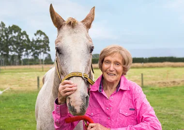 Senior woman in pink shirt smiling beside a white horse in a field.