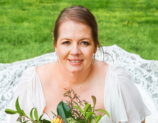 Bride in white dress holding a bouquet, smiling, sitting on grass.