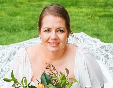 Bride in white dress holding a bouquet, smiling, sitting on grass.