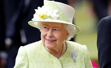 A smiling elderly woman in a light green outfit and hat adorned with white flowers, wearing a pearl necklace.