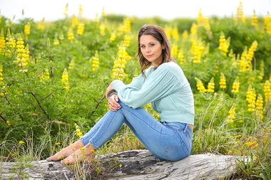 Woman in light blue sweater and jeans sitting on a log amidst yellow flowers and greenery.
