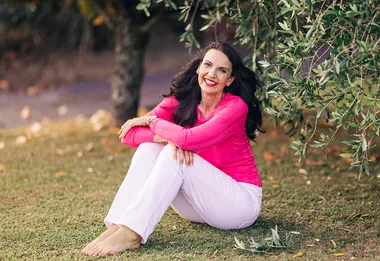 A woman in a pink top and white pants sits barefoot on grass near a leafy tree, smiling.